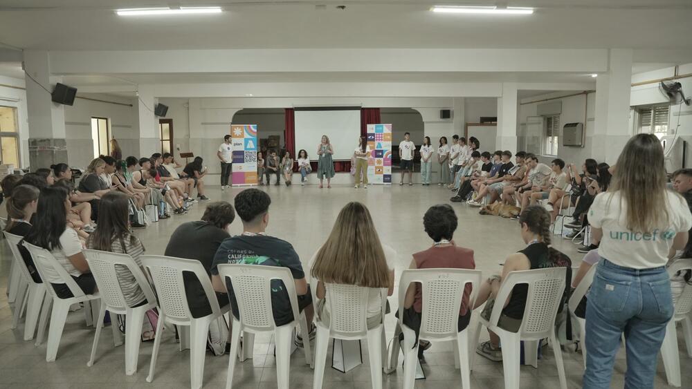 Grupo numeroso de adolescentes y jóvenes sentados en sillas en un salón amplio. Otras personas hablan de pie