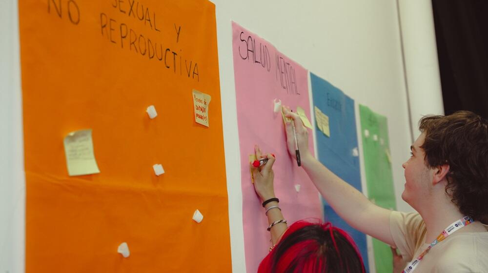 Adolescentes y jóvenes pegando papeles sobre afiches colgados en una pared.